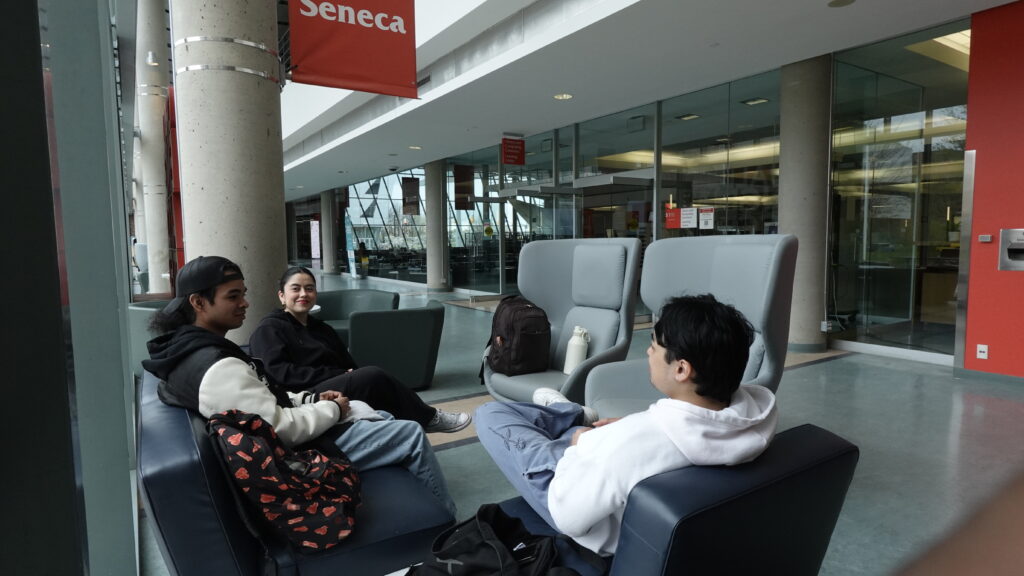 International students relaxing and talking in a modern campus lounge under the Seneca logo at Seneca Polytechnic.